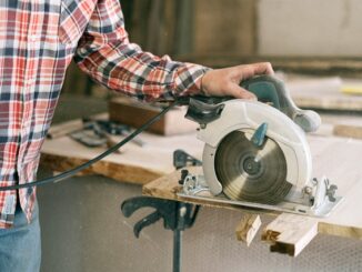 Na zdjęciu widzimy postać tnącą drzewo stołową piłą tarczową. Osoba jest ubrana w koszule w kratę i jeansy. Piła jest w kolorze biało-zielonym. Na ilustracji widać czarne zaciski mocujące piłę do drewnianego stołu.