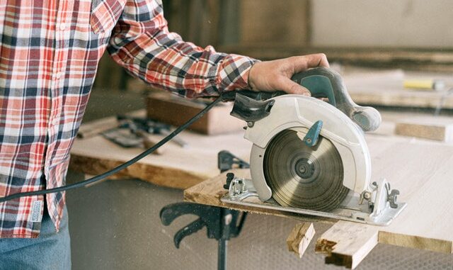 Na zdjęciu widzimy postać tnącą drzewo stołową piłą tarczową. Osoba jest ubrana w koszule w kratę i jeansy. Piła jest w kolorze biało-zielonym. Na ilustracji widać czarne zaciski mocujące piłę do drewnianego stołu.