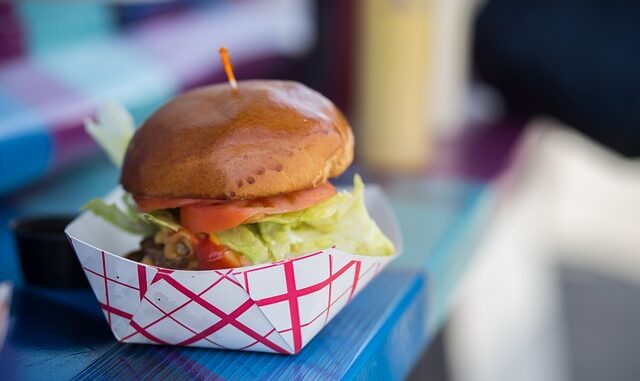 Na grafice widzimy burgera w opakowaniu w biało-czerwoną kratkę. Kanapka składa się z bułki, pomidora, sałaty, sosu i kotleta. Całość burgera jest spięta wykałaczką.
