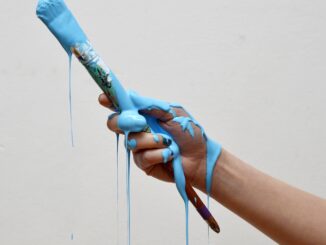 Grafika przedstawia dłoń trzymająca pędzel. Z pędzla i dłoni ocieka farba w kolorze niebieskim.