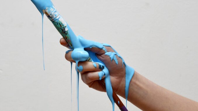 Grafika przedstawia dłoń trzymająca pędzel. Z pędzla i dłoni ocieka farba w kolorze niebieskim.