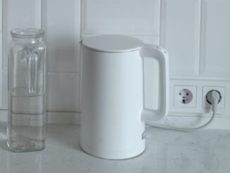 W centralnym punkcie zdjęcia znajduje się biały czajnik podłączony białym kablem do jednego z dwóch gniazd sieciowych. Po lewej stronie czajnika znajduje się przezroczysty dzbanek na wodę.