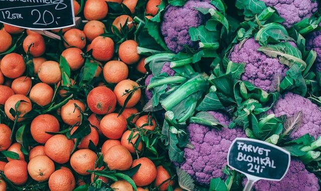 Na grafice widzimy stragan z mandarynkami i fioletowymi kalafiorami. Na zdjęciu widać czarne tabliczki z cenami owoców i warzyw.