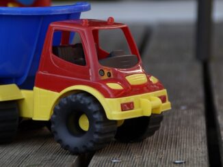 Na zdjęciu znajduje widzimy zabawkową ciężarówkę. Kabina ciężarówki jest w kolorze czerwonym, zderzaki, podwozie, felgi, błotniki i światła są w kolorze żółtym, natomiast przyczepa jest w barwie niebieskiej. Ciężarówka stoi na drewnianym podłożu w kolorze brązowym.
