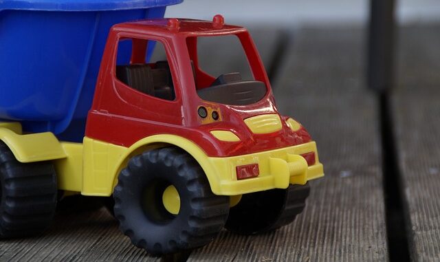 Na zdjęciu znajduje widzimy zabawkową ciężarówkę. Kabina ciężarówki jest w kolorze czerwonym, zderzaki, podwozie, felgi, błotniki i światła są w kolorze żółtym, natomiast przyczepa jest w barwie niebieskiej. Ciężarówka stoi na drewnianym podłożu w kolorze brązowym.