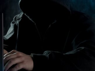 Grafika przedstawia postać siedzącą w czarnej bluzie z założonym kapturem na głowę. Nie widać twarzy osoby przedstawionej na zdjęciu. Dłonie postaci położone są na klawiaturze laptopa. Klapa monitora jest w kolorze czarnym.