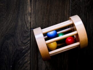 Grafika przedstawia drewnianą grzechotkę w kształcie walca. W środku zabawki znajdują się cztery kulki w kolorach żółtym, czerwonym, zielonym i niebieskim. Zabawka leży na drewnianym podłożu o barwie ciemnego brązu.