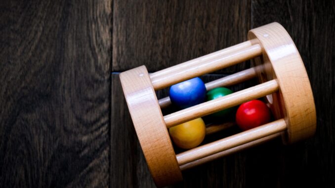 Grafika przedstawia drewnianą grzechotkę w kształcie walca. W środku zabawki znajdują się cztery kulki w kolorach żółtym, czerwonym, zielonym i niebieskim. Zabawka leży na drewnianym podłożu o barwie ciemnego brązu.