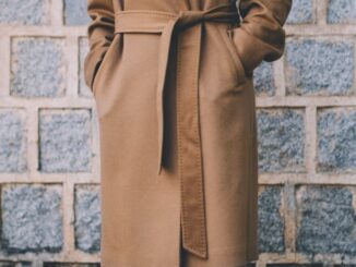 Na centralnej części obrazka widzimy postać w długim beżowym płaszczu, przepasanym paskiem. Osoba na zdjęciu ma włożone dłonie do kieszeni płaszcza. Tło obrazka stanowią szare pustaki z jasną fugą.