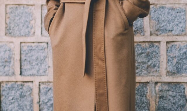 Na centralnej części obrazka widzimy postać w długim beżowym płaszczu, przepasanym paskiem. Osoba na zdjęciu ma włożone dłonie do kieszeni płaszcza. Tło obrazka stanowią szare pustaki z jasną fugą.